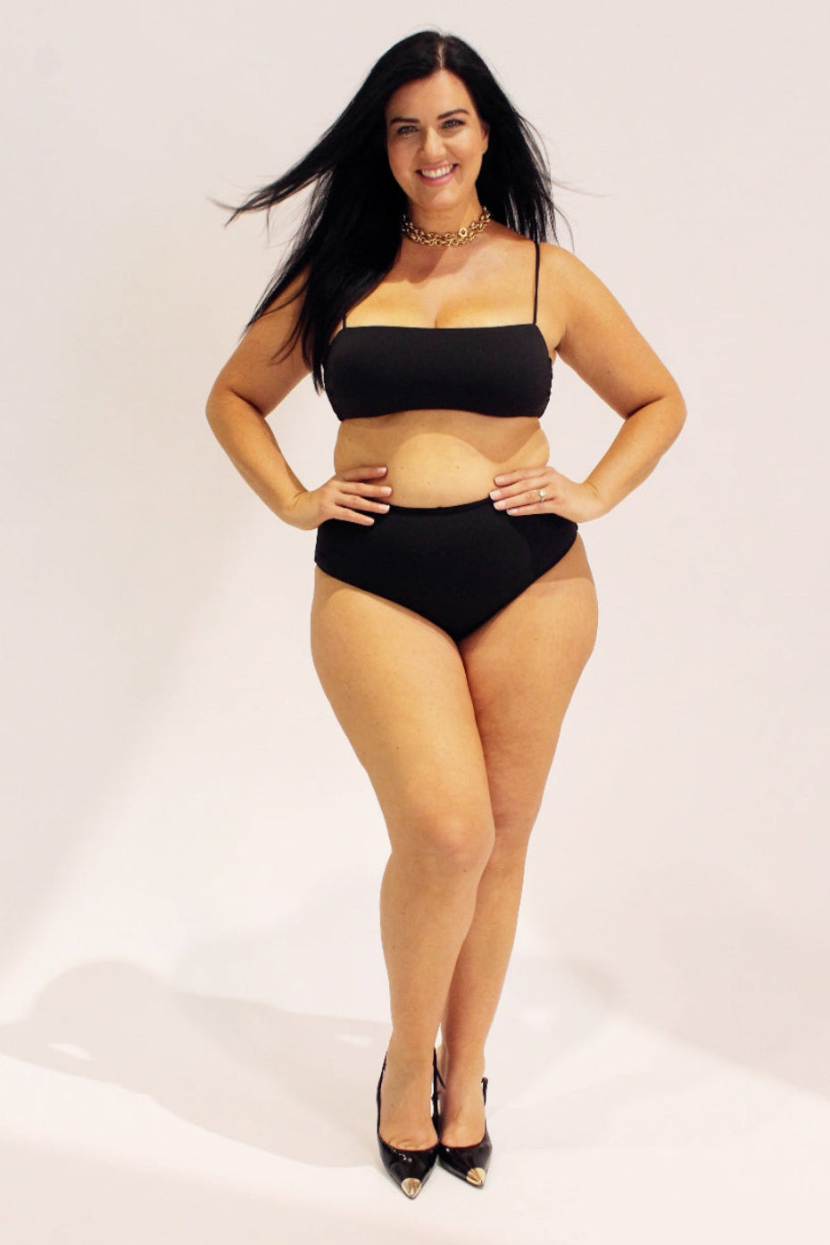A woman modeling a black two-piece bikini set with high-waisted bottoms and thin shoulder straps, paired with black pointed-toe heels, against a plain white studio background.